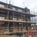 A man is standing in front of a building with scaffolding, possibly working on loft insulation.