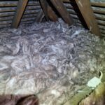 Thick layer of fiberglass insulation installed between wooden beams in an attic, with visible wiring and part of a ladder.