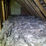 Dusty attic filled with thick layers of insulation; wooden beams and a chimney structure are visible.