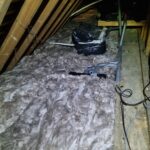 An attic space with thick insulation, a taped plastic bag, and scattered tools on the floor, with exposed wooden beams and pipes visible.