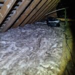 Insulation material spread across an attic floor, with exposed wooden beams and a tool bag, lit dimly.