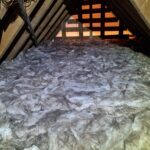 Insulation material filling the floor of an attic, with wooden beams and a dim light in the background.