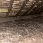 Dusty attic space with exposed wooden beams and insulation, featuring a visible brick wall and a partial wooden stair edge.