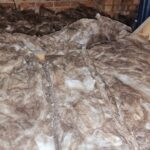 Insulation material spread across an attic floor, featuring wood beams and partial view of a roof structure.