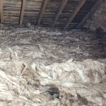 Loose fiberglass insulation covering the floor of an unfinished attic with exposed wooden beams and brick walls.