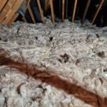 Insulation material covers the floor in an attic space, surrounded by wooden beams, with exposed wires and debris visible.