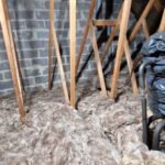 An attic filled with insulation material, with exposed wooden beams and a bag covered in black plastic.