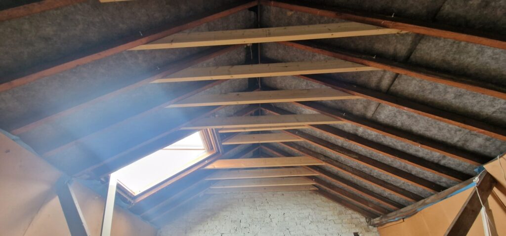 Interior view of an unfinished attic with exposed wooden beams and insulation, featuring a skylight and white brick walls.