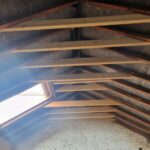 Interior view of an unfinished attic with exposed wooden beams and insulation, featuring a skylight and white brick walls.