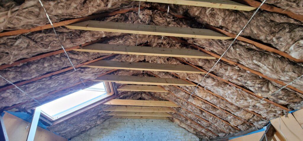 Interior of an unfinished attic with exposed insulation between wooden beams and a skylight.
