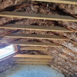 Interior of an unfinished attic with exposed insulation between wooden beams and a skylight.