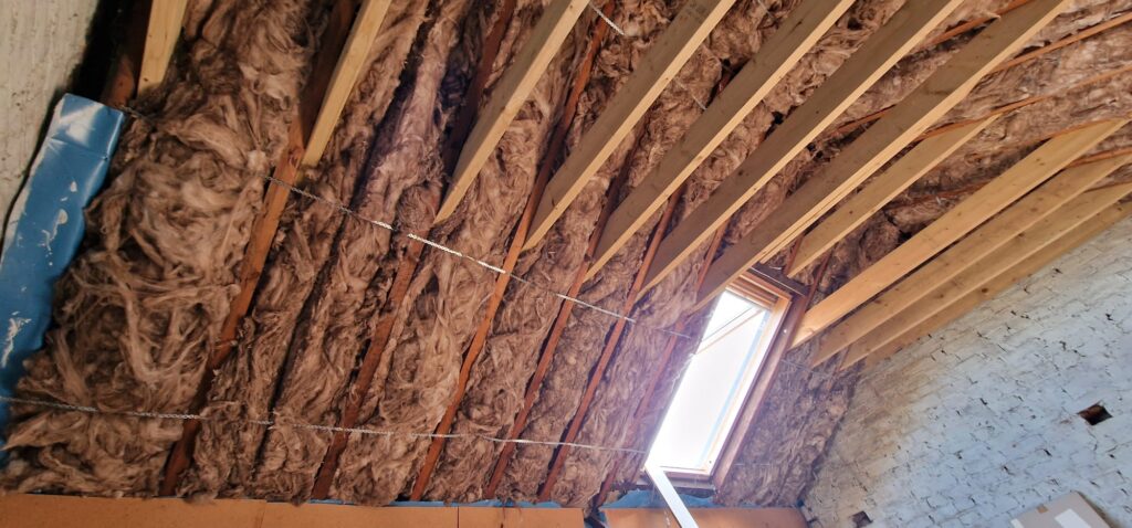 Interior view of a house under construction showing exposed wooden beams and pink fiberglass insulation material on the ceiling, with a small window letting in light.