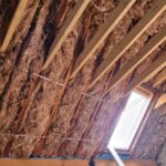 Interior view of a house under construction showing exposed wooden beams and pink fiberglass insulation material on the ceiling, with a small window letting in light.