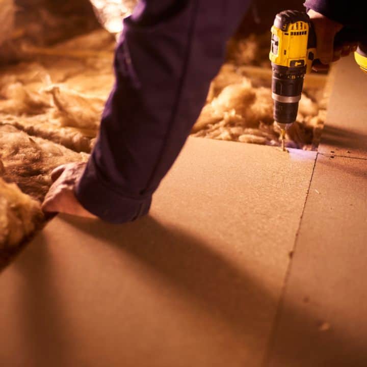 Person installing loft boarding with power drill and insulation material.