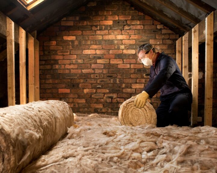 A man is working on a roll of insulation in an attic, installing loft insulation.